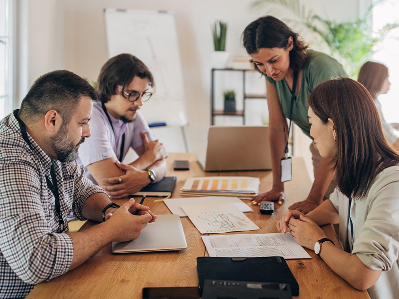 A diverse group of 4 people in a meeting with papers and laptops on a wood table.